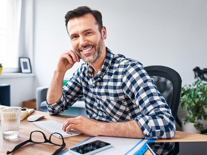A man in a black and white checkered button-up sitting at a desk and smiling